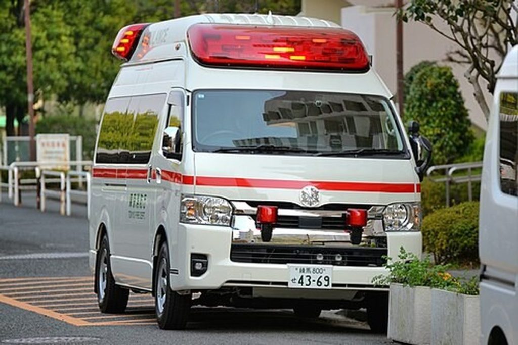 White Japanese ambulance with a red stripe and red roof lights parked outside an apartment building
