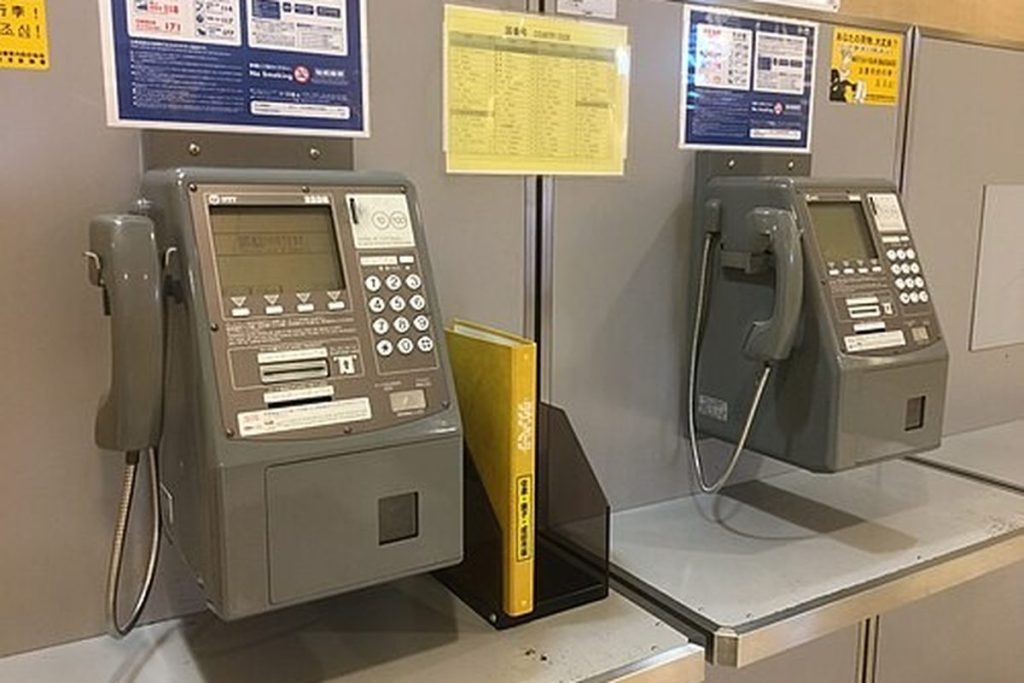 Two grey digital public telephones on a counter at Narita Airport with instruction posters on the wall