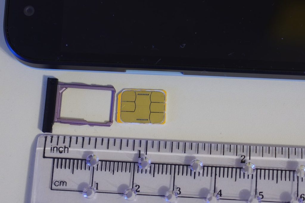 A nano SIM card next to an empty phone SIM tray and a ruler, showing the small card a physical SIM option needs