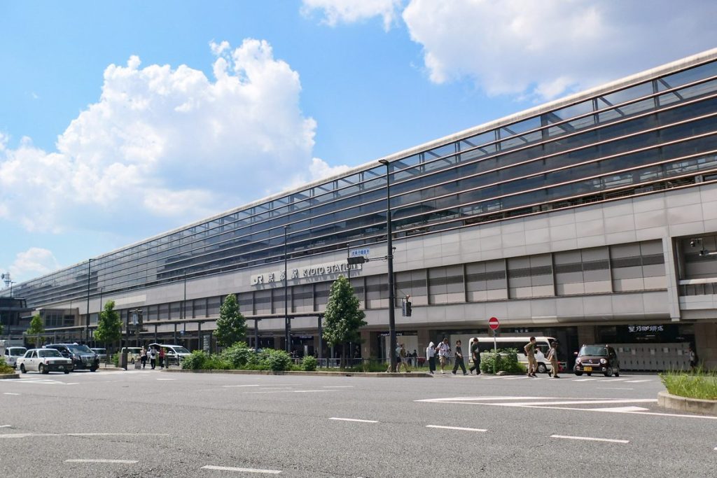 Long modern facade of the Hachijo exit on the south side of Kyoto Station, with taxis and cars along the road under a blue sky