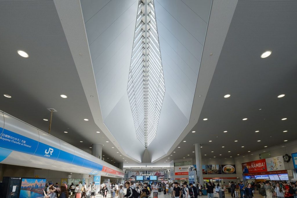 Busy concourse of Kansai Airport Station under a tall glass roof, with the blue JR West ticket gates on the left and the Nankai side further back