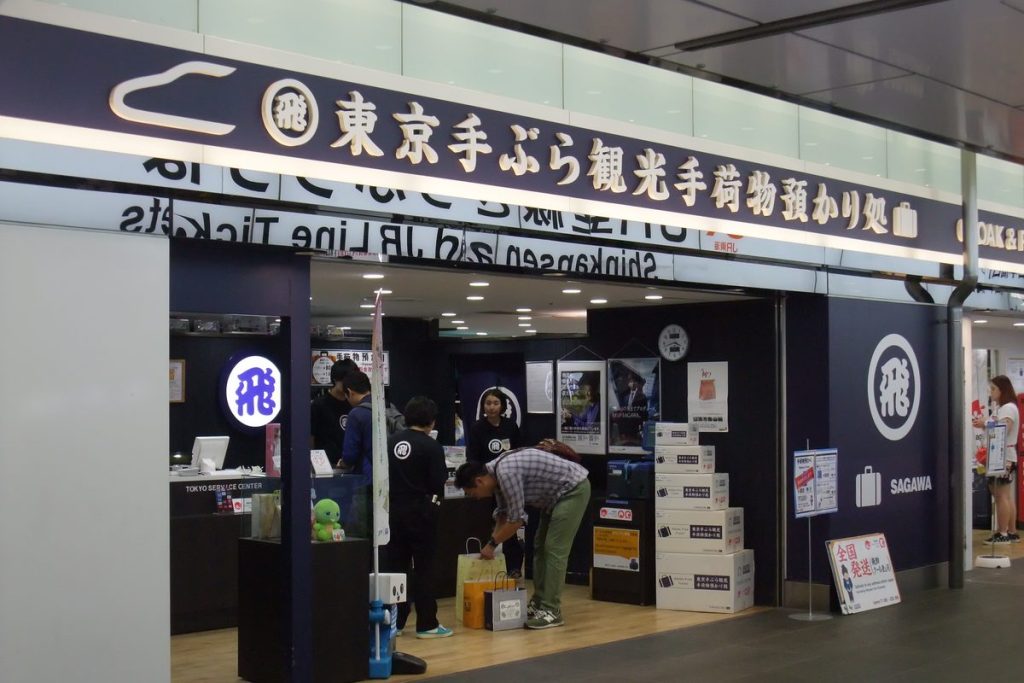 Hands-free travel baggage counter in Tokyo Station's First Avenue mall, with a large Japanese sign above the entrance, staff at the counter and stacked boxes inside