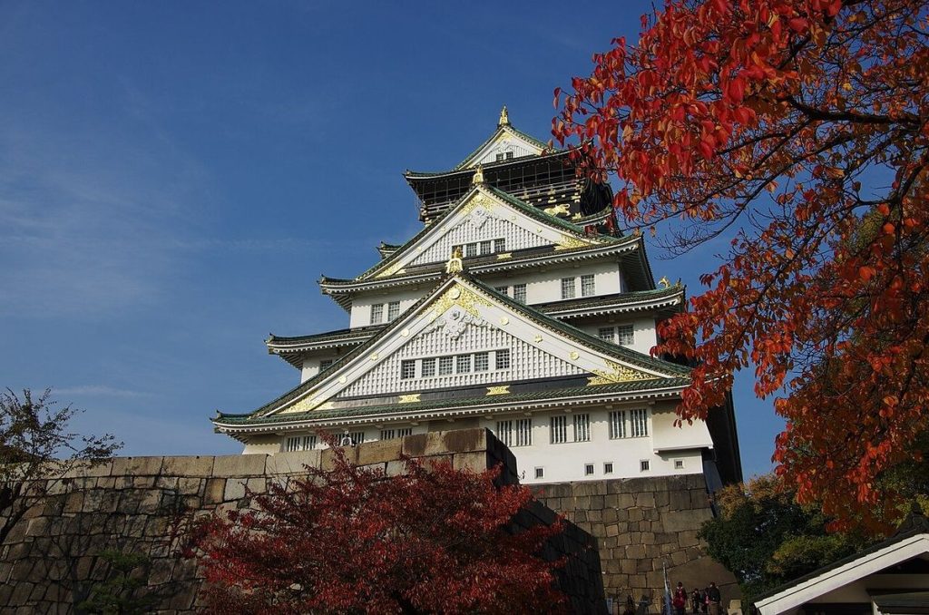 Osaka Castle's white and green main tower above its stone walls, framed by red autumn maple leaves under a blue sky