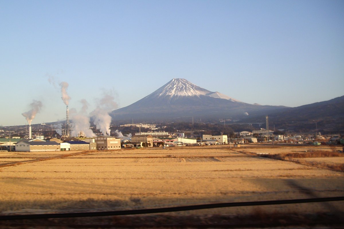 Mount Fuji seen across fields and factory chimneys from the window of a Tokaido Shinkansen train