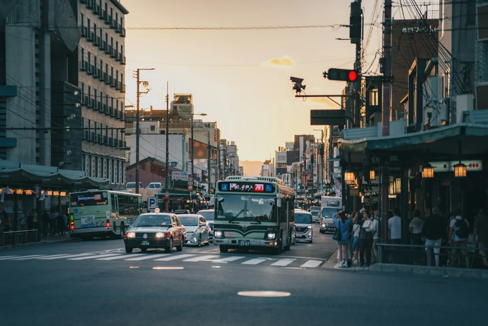 A light-green Kyoto City Bus waiting in traffic beside a taxi on a busy central Kyoto street at sunset